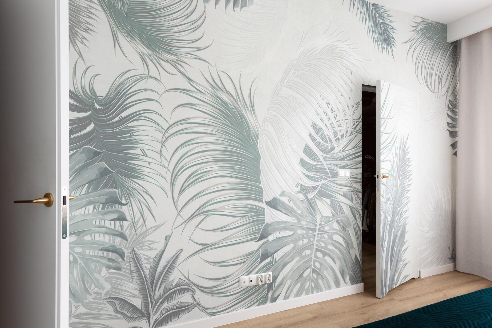 Wnętrze pokoju z tapetą w motywy tropikalnych liści, widoczna biała ościeżnica drzwiowa z złotą klamką i fragment drewnianej podłogi.