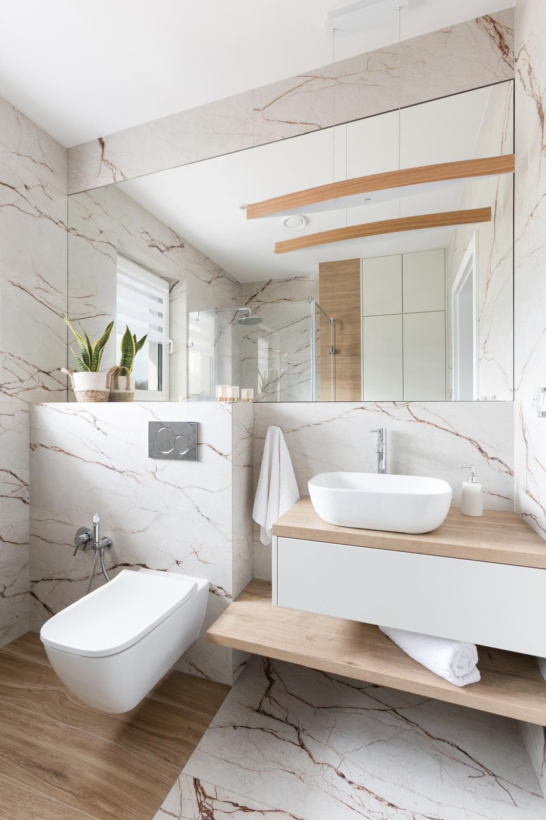 Nowoczesna łazienka z marmurowymi płytkami, podwieszaną toaletą, owalną umywalką na drewnianym blacie i dużym lustrem odbijającym kabinę prysznicową.