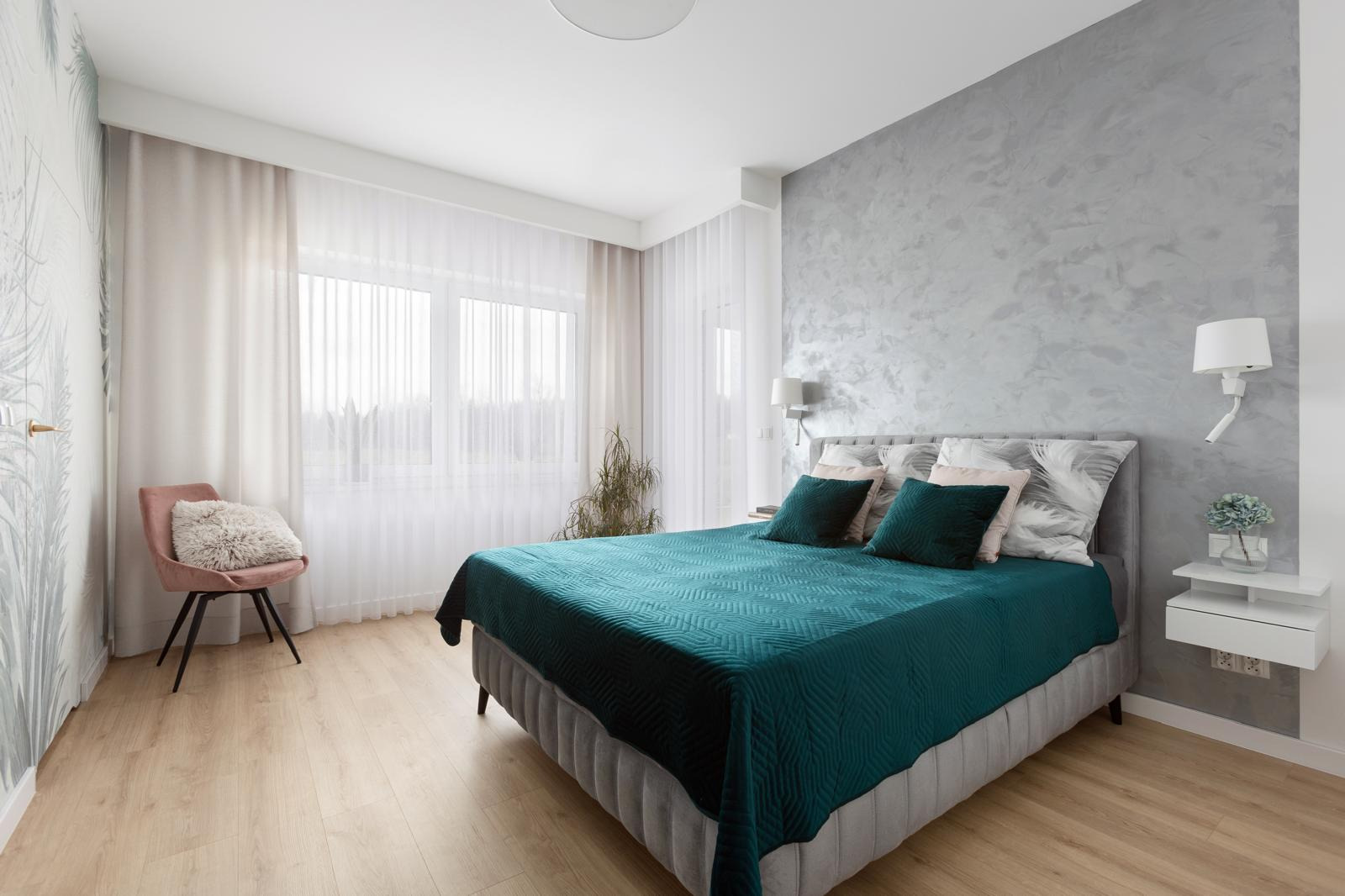 Jasna sypialnia z łóżkiem kontynentalnym, turkusową narzutą, szarą ścianą z efektem dekoracyjnym, białymi zasłonami i akcentem w postaci pudrowego fotela z poduszką.