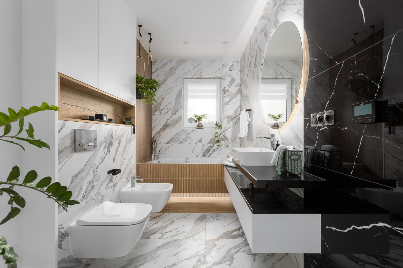 Elegancka łazienka z marmurowymi płytkami, białymi meblami i czarnym blatem, wyposażona w wannę, umywalkę, bidet i toaletę podwieszaną, z oświetlonym lustrem owalnym i roślinami wiszącymi.