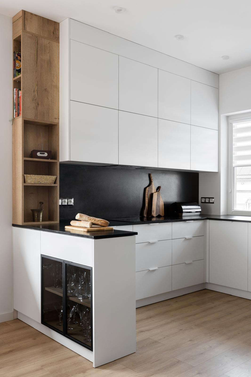Nowoczesna kuchnia z białymi szafkami, czarnym blatem i drewnianym regałem, na blacie chleb na desce i akcesoria kuchenne.