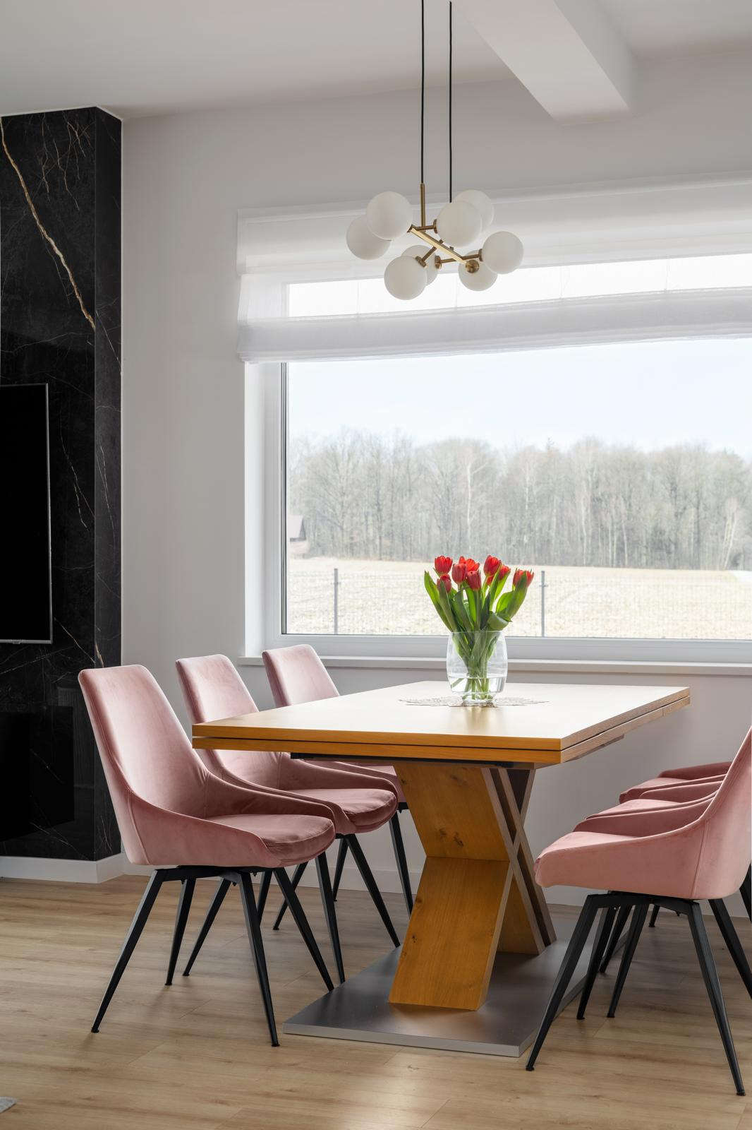 Jasna jadalnia z drewnianym stołem na metalowej podstawie, otoczonym różowymi krzesłami z czarnymi nogami, z widokiem na krajobraz za oknem i nowoczesną lampą sufitową.