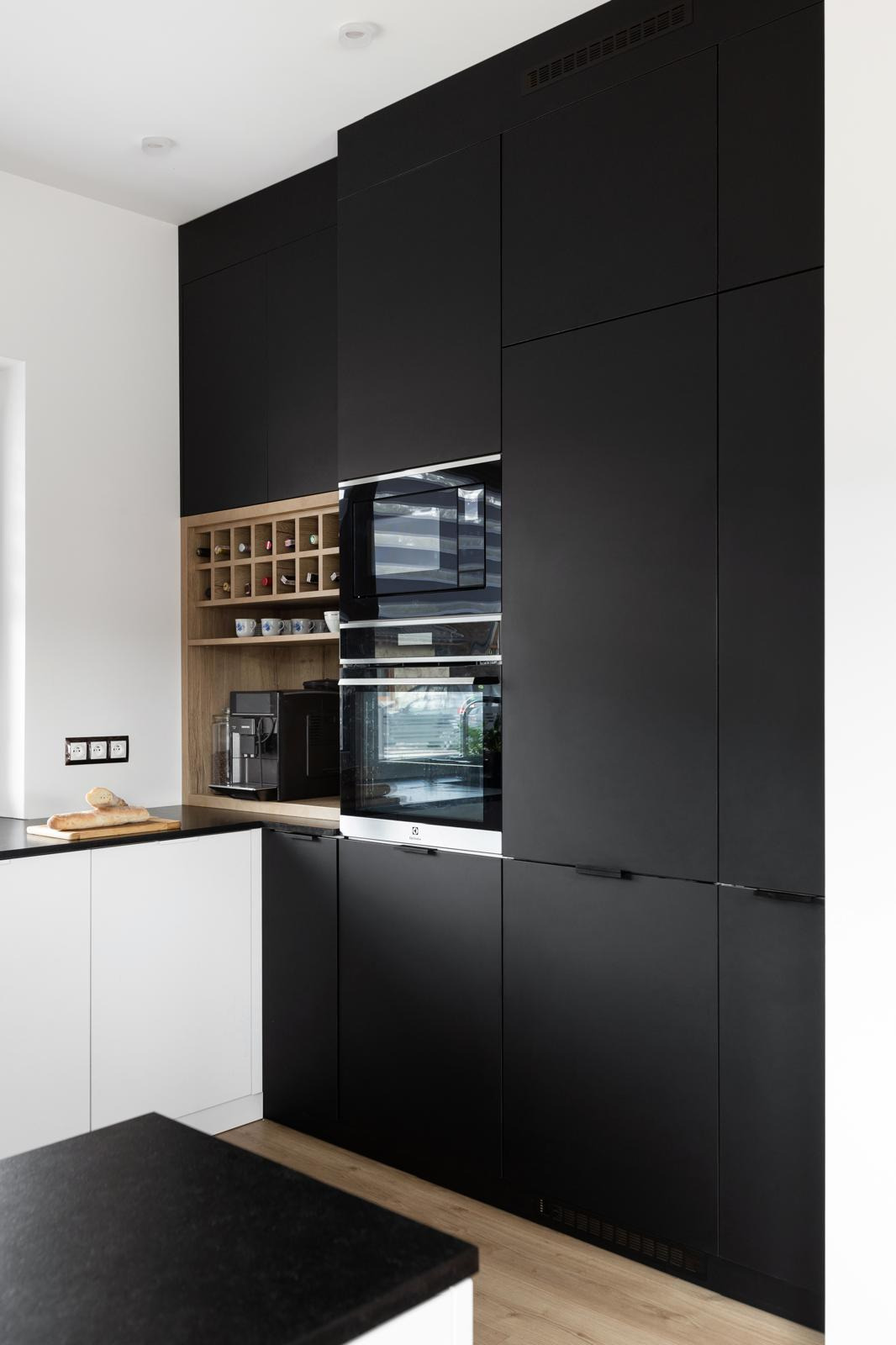 Nowoczesna kuchnia z czarnymi szafkami, drewnianym regałem na wino, ekspresem do kawy i wbudowanym piekarnikiem.