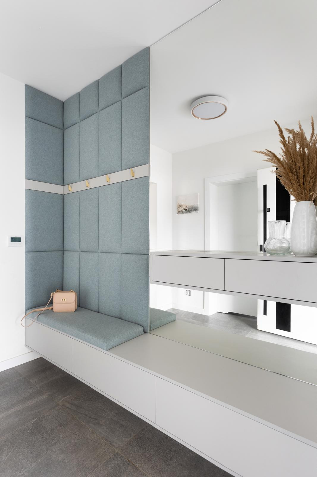 Wnęka w przedpokoju z siedziskiem obitym jasnoniebieską tkaniną, panelami ściennymi w tym samym kolorze i złotymi haczykami; na siedzisku leży mała, beżowa torebka.