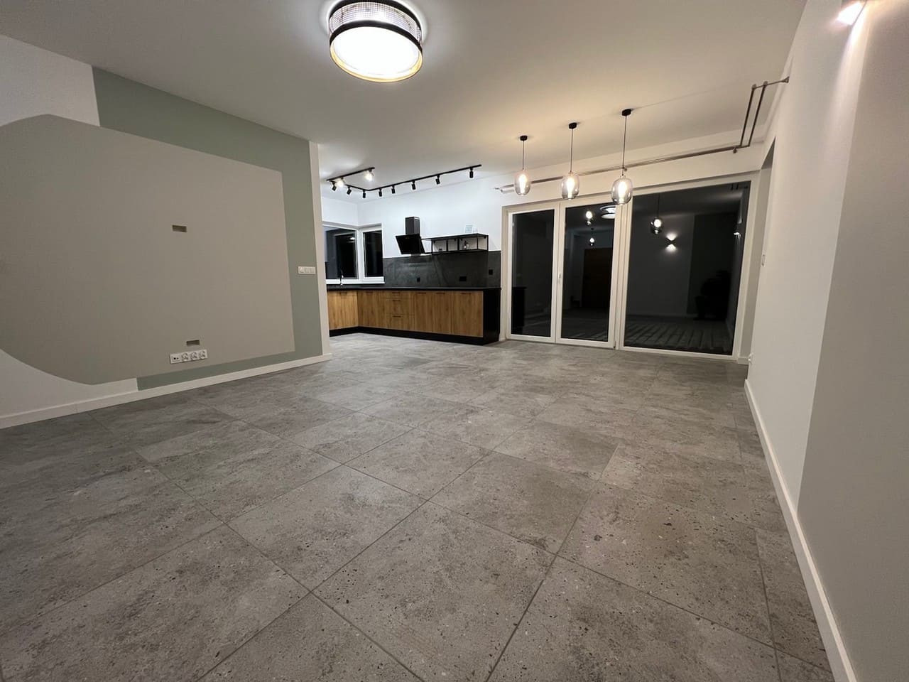 Nowoczesne wnętrze domu z szarą podłogą, drewnianą zabudową kuchenną i dużymi przesuwnymi drzwiami tarasowymi, oświetlone punktowymi lampami sufitowymi.