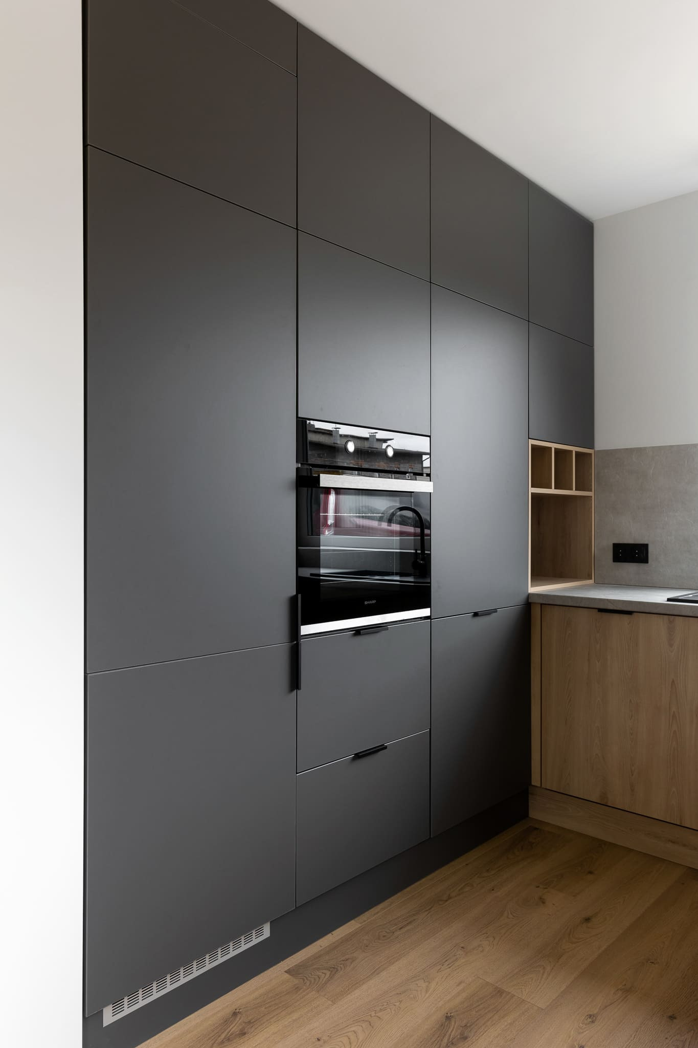 Nowoczesna kuchnia z zabudową w odcieniach szarości i drewna, z piekarnikiem w zabudowie i drewnianą podłogą.