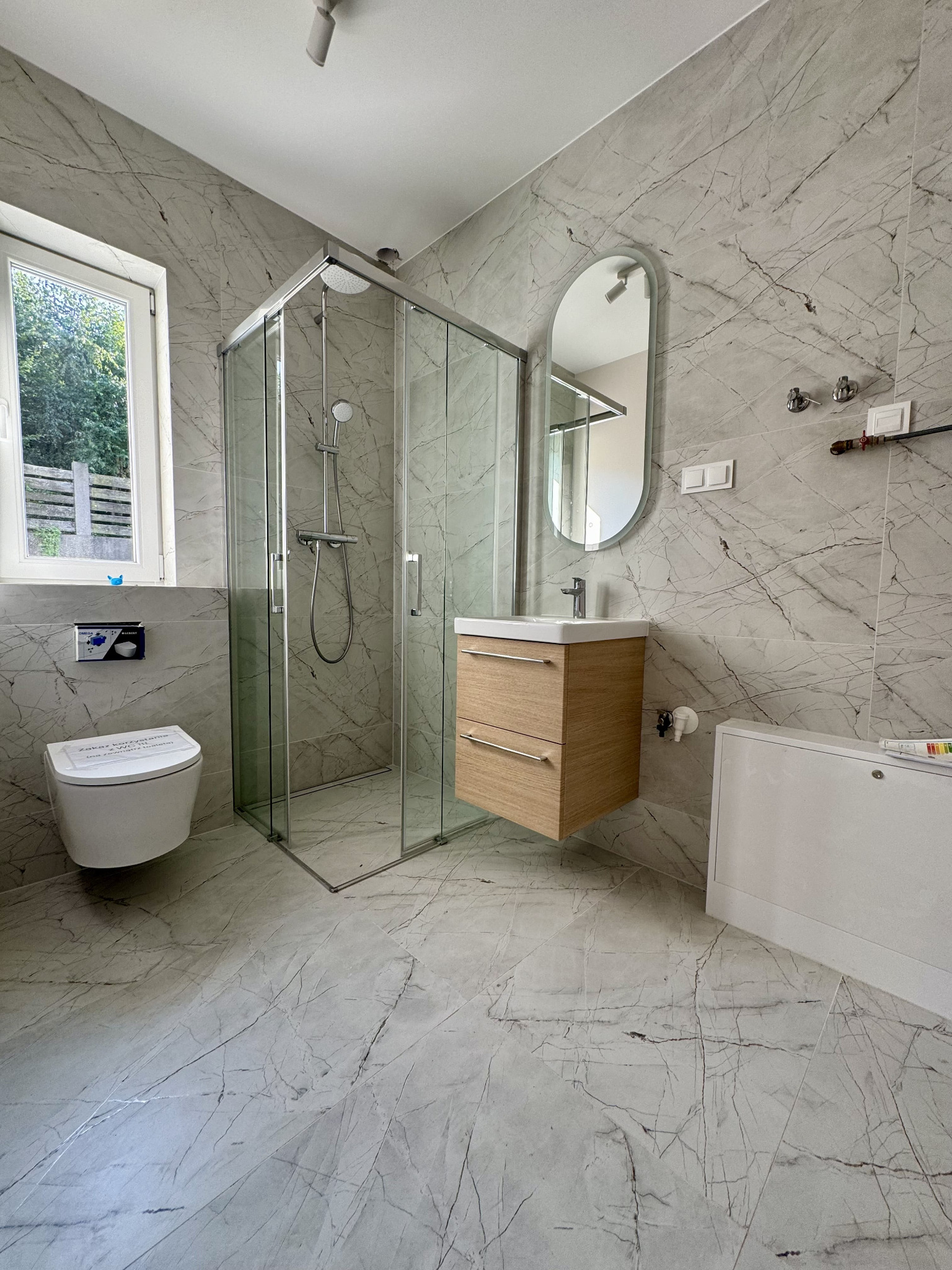 Nowoczesna łazienka z marmurowymi płytkami, kabiną prysznicową ze szkła, podwieszaną szafką z umywalką, owalnym lustrem i podwieszaną toaletą z niebieskim przyciskiem spłukiwania.