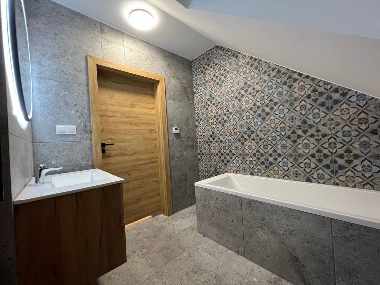 Nowoczesna łazienka z drewnianymi drzwiami i wzorzystą ścianą nad wanną, oświetlona lampą sufitową.