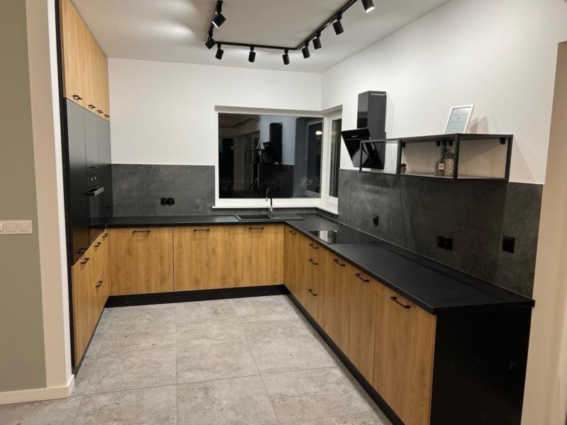 Nowoczesna kuchnia z drewnianymi frontami szafek, czarnym blatem i szarą ścianą nad blatem, oświetlona reflektorami sufitowymi.