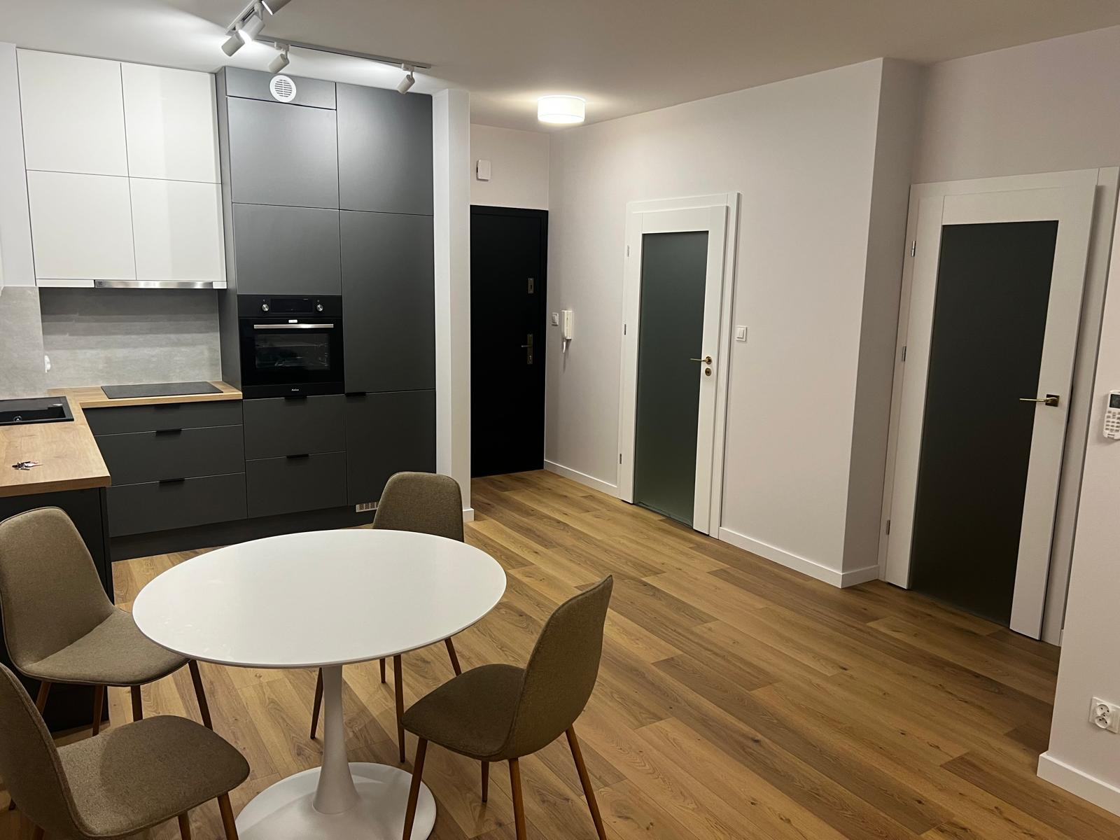 Nowoczesna kuchnia z szarymi szafkami, drewnianym blatem i okrągłym białym stołem z czterema krzesłami na drewnianej podłodze. Widoczne trzy drzwi wewnętrzne z ciemnymi szybami.