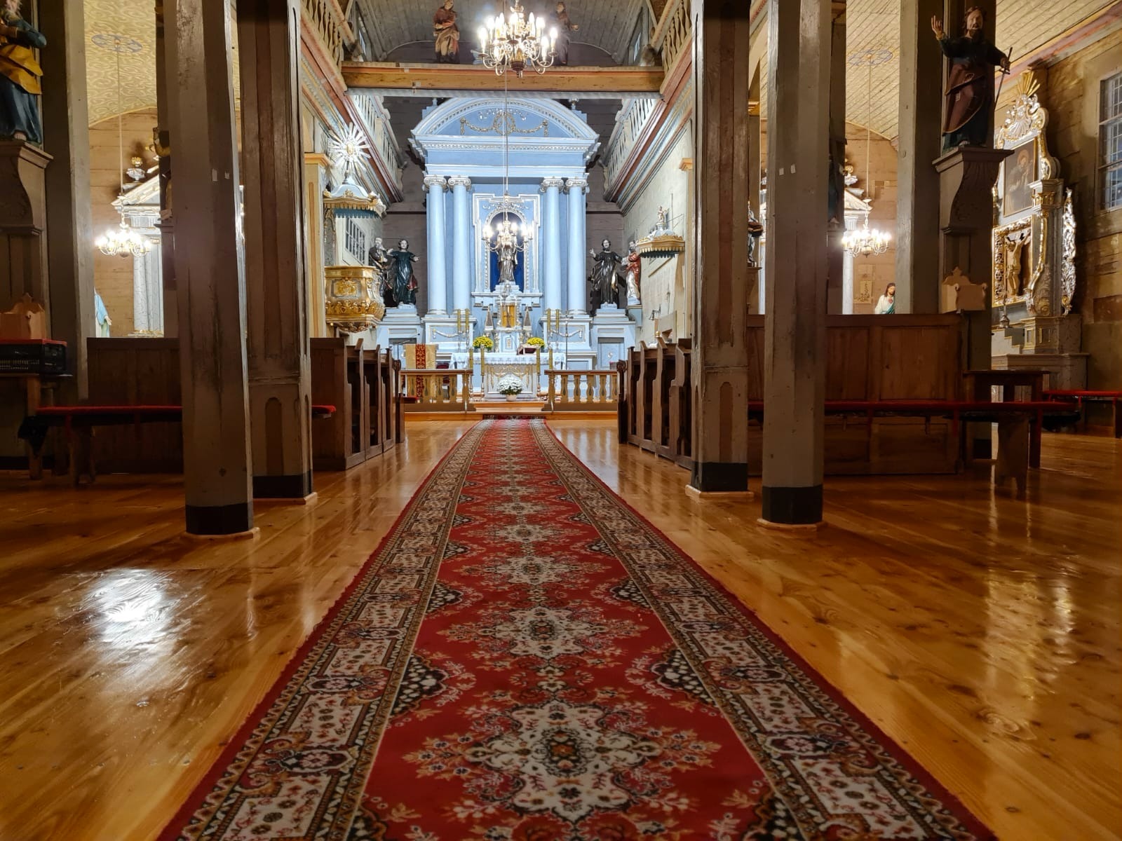 Wnętrze drewnianego kościoła z czerwonym dywanem prowadzącym do ołtarza, widoczne drewniane ławki i kolumny, oświetlone żyrandolami i światłem naturalnym.
