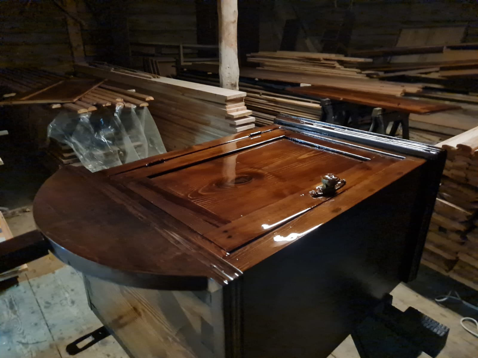 Odnowiona szafka z drewna sosnowego z błyszczącą, brązową politurą, widoczny metalowy uchwyt i fragment półokrągłego blatu, w tle stosy surowego drewna.