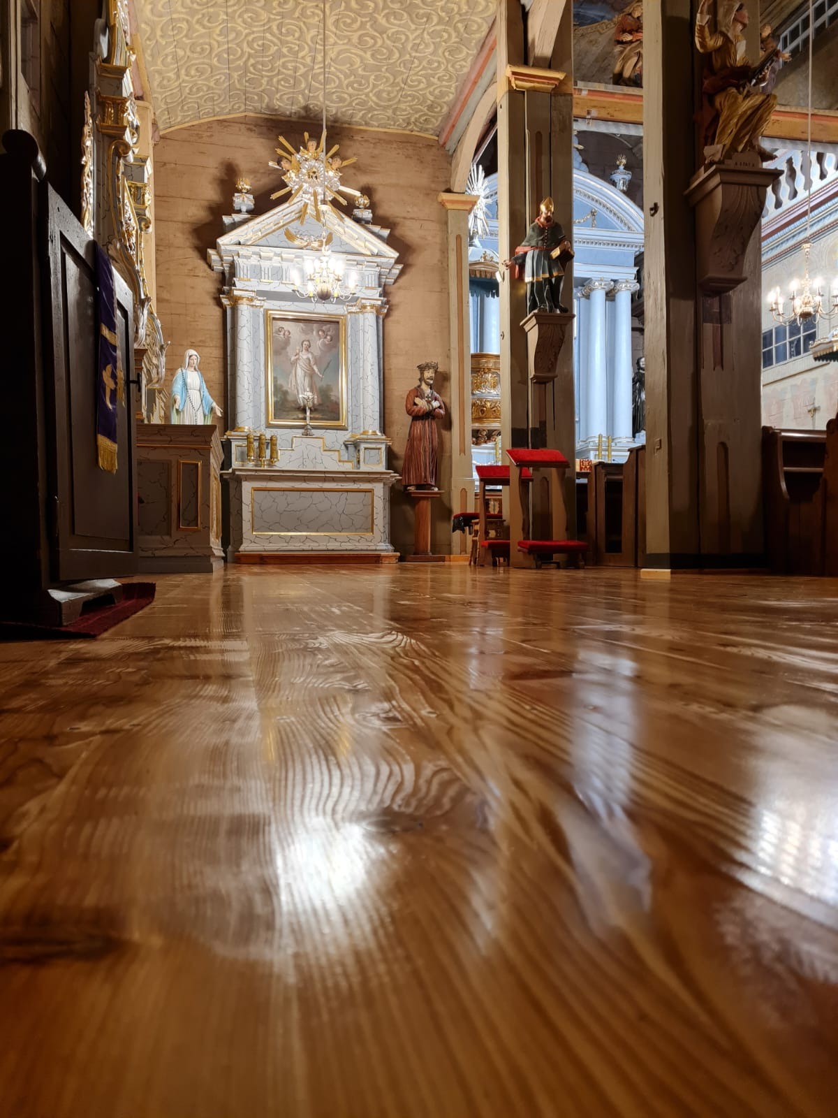 Wnętrze zabytkowego kościoła z drewnianą podłogą odbijającą światło, widok na ołtarz z obrazem, figury świętych i kolumny.