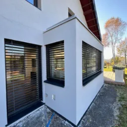 Elewacja domu z trzema oknami zabezpieczonymi poziomymi, metalowymi osłonami przeciwsłonecznymi w kolorze czarnym, widoczne z zewnątrz w jesiennym świetle.