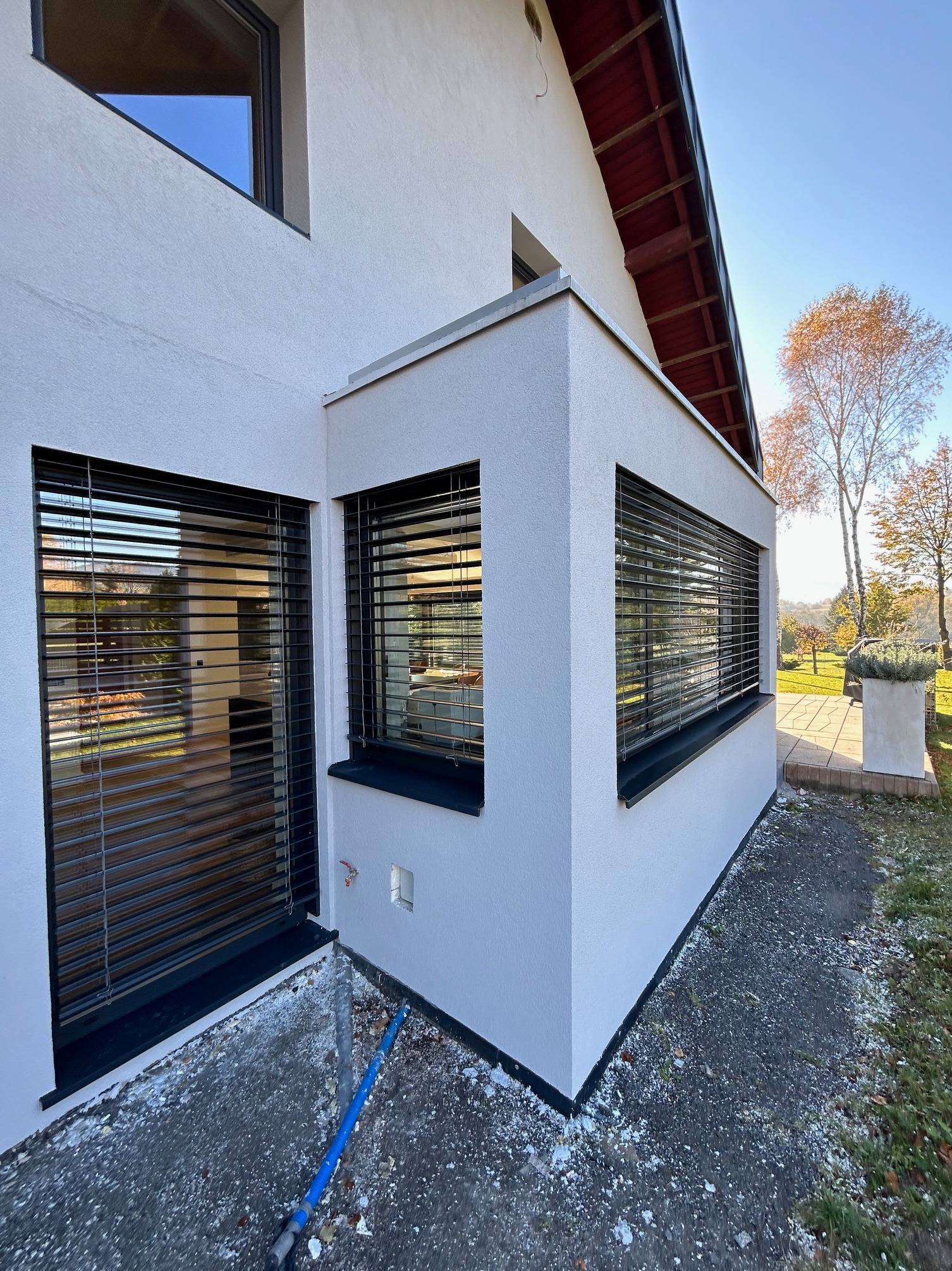 Elewacja domu z trzema oknami zabezpieczonymi poziomymi, metalowymi osłonami przeciwsłonecznymi w kolorze czarnym, widoczne z zewnątrz w jesiennym świetle.