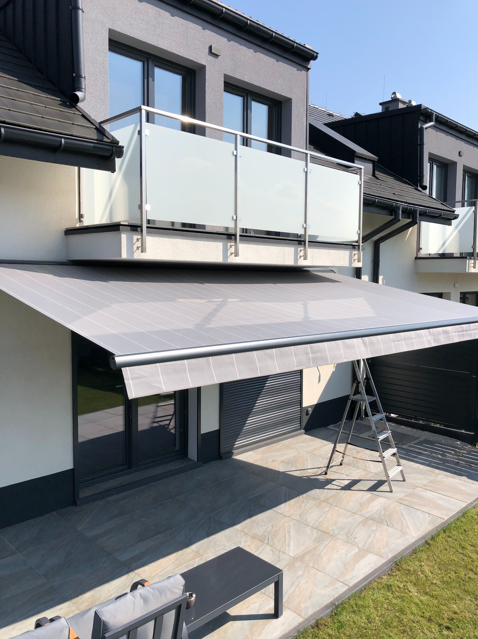 Szara, rozłożona osłona przeciwsłoneczna zamontowana nad tarasem nowoczesnego domu z balkonem i szklaną balustradą, drabina oparta o ścianę.