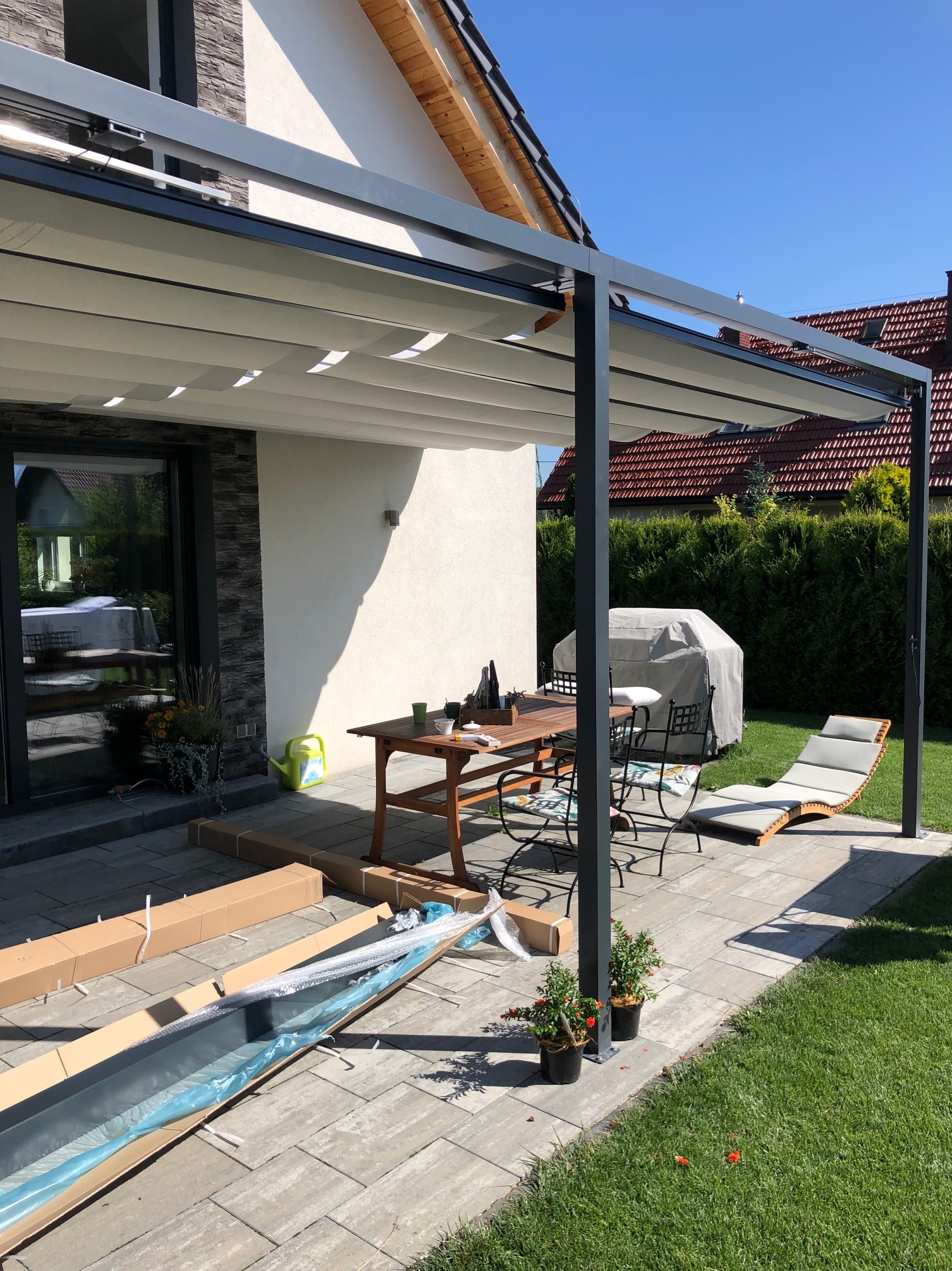 Montaż pergoli tarasowej z rozsuwanym zadaszeniem, widoczne rozpakowane elementy konstrukcji na kostce brukowej, stół z krzesłami i leżak na trawniku, w tle dom z ogrodem.