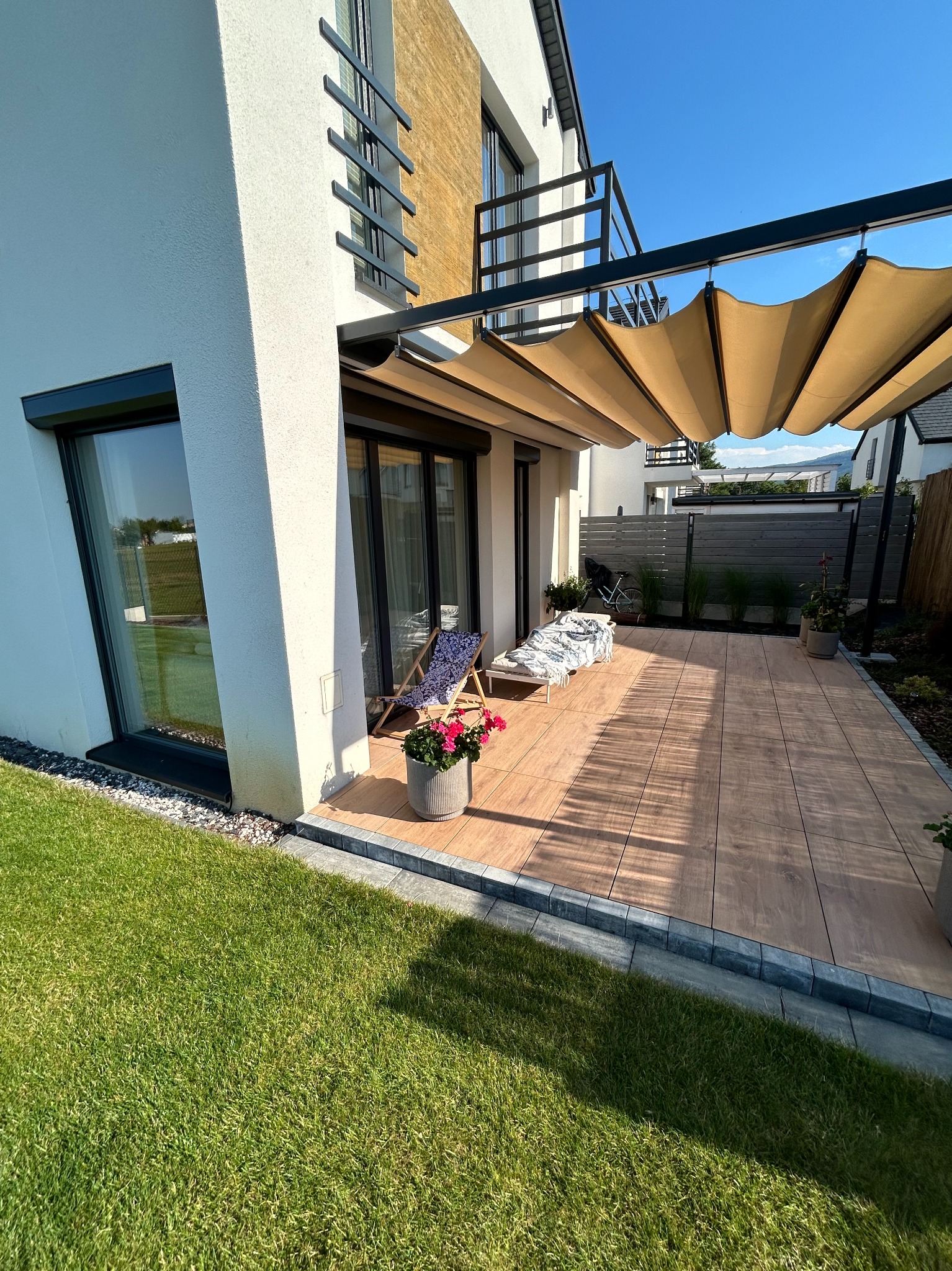 Nowoczesna pergola z beżową, zwijaną tkaniną nad tarasem z drewnopodobnych płytek, leżaki z niebieskim wzorem, kwiaty w szarej donicy, trawnik, biały dom z czarnymi ramami okiennymi i balkonem.
