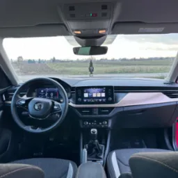 Wnętrze samochodu Skoda z widokiem na deskę rozdzielczą, kierownicę i przednią szybę z zawieszką zapachową. Na zewnątrz widoczny krajobraz polny.