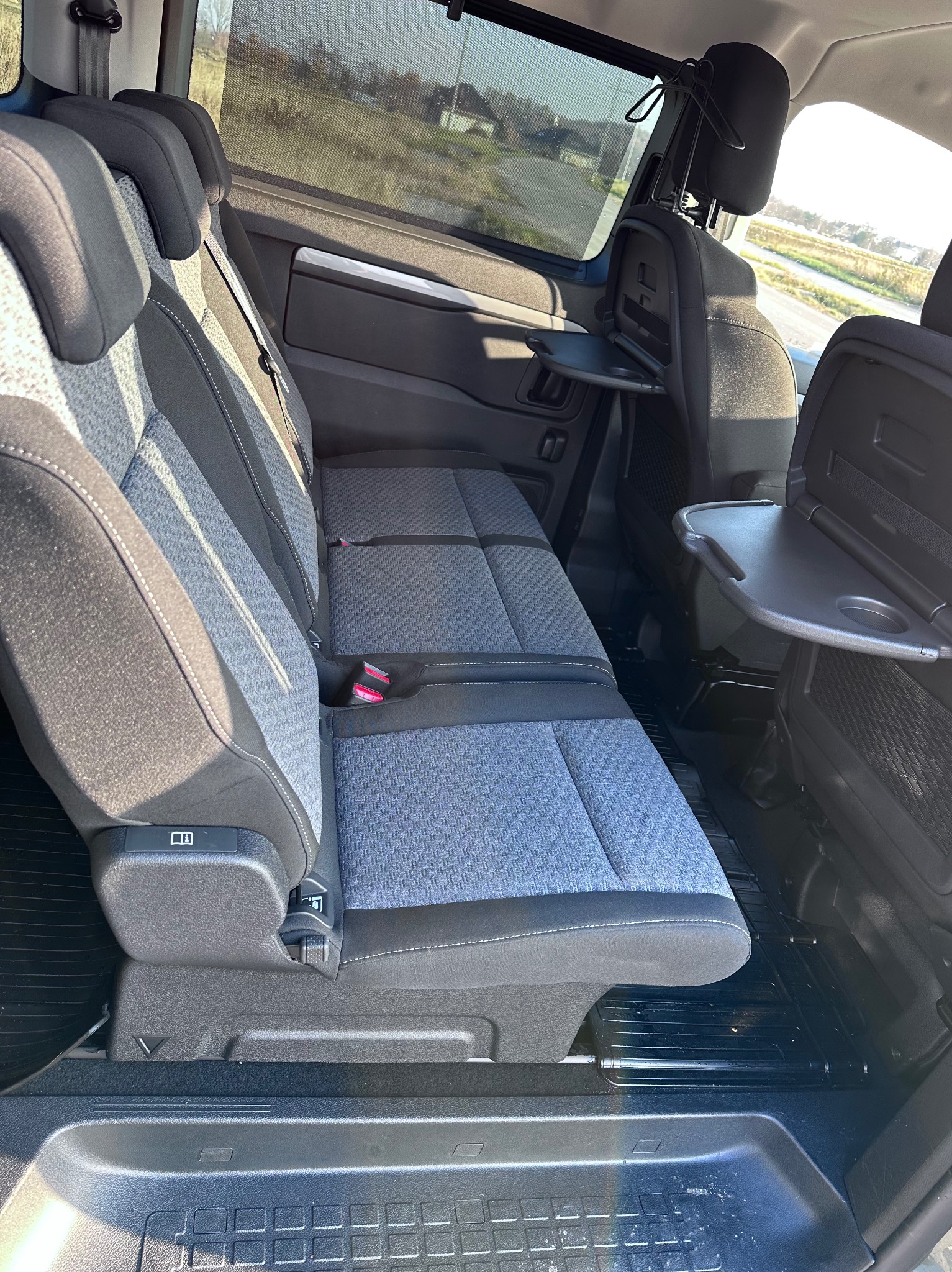 Wnętrze busa z trzema rzędami siedzeń w odcieniach szarości i czerni, rozkładane stoliki przymocowane do oparć przednich foteli, widok z perspektywy pasażera.