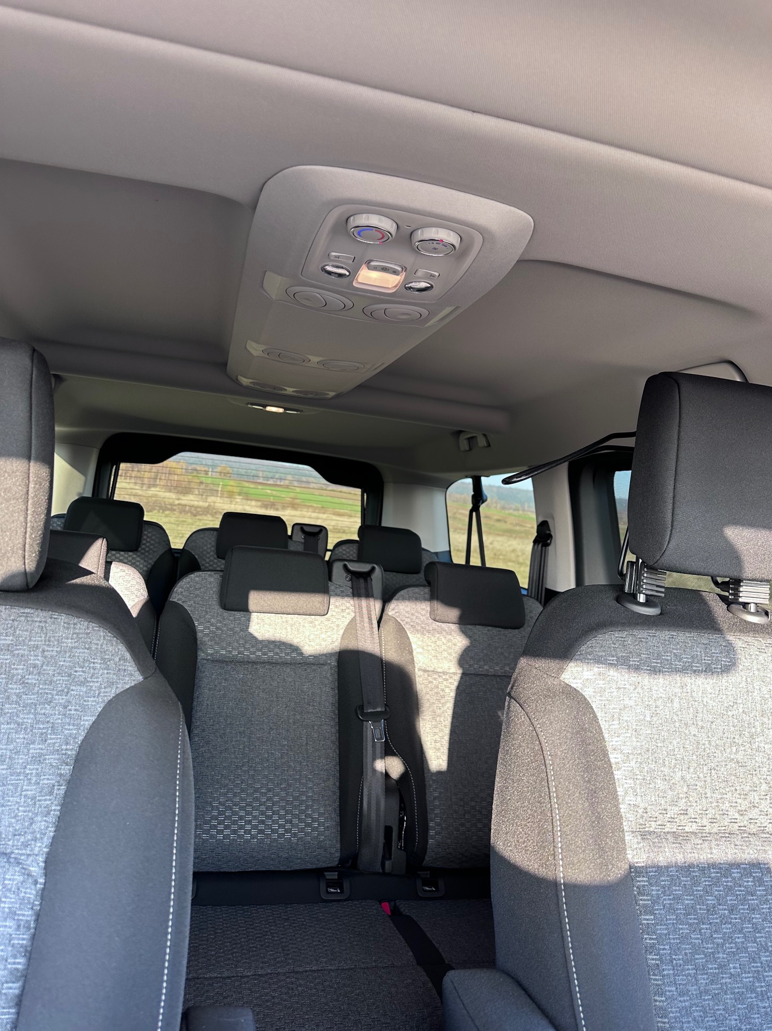 Wnętrze przestronnego busa z szarą tapicerką, widok z przodu na tylne siedzenia, zagłówki i system oświetlenia sufitowego z regulacją, za oknem krajobraz polny.