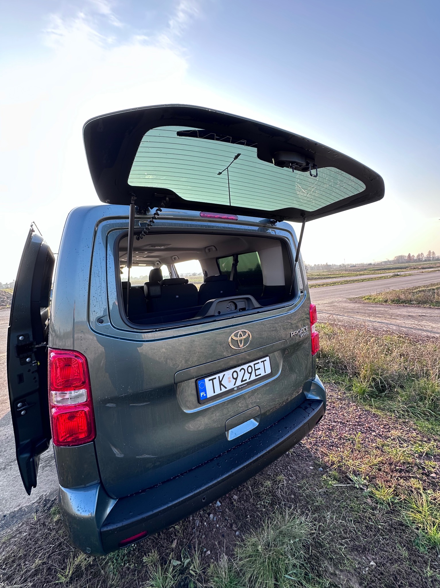 Zielony van Toyota Proace Verso z otwartą klapą bagażnika, widok z tyłu na tle polnej drogi i zachodzącego słońca.