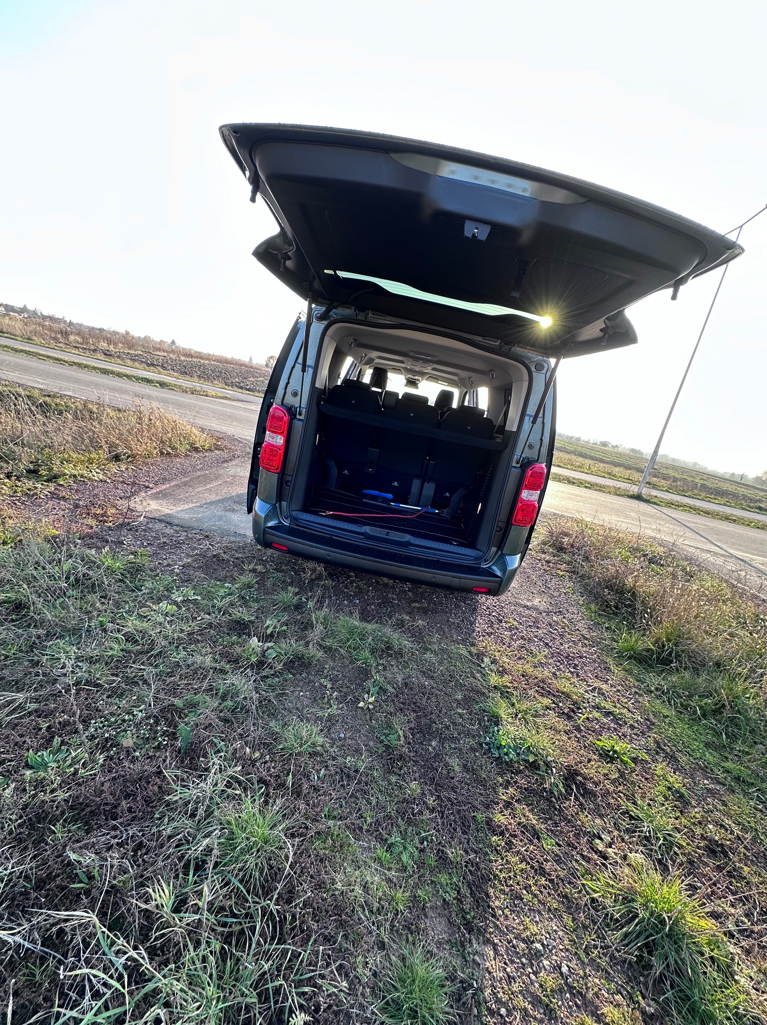 Otwarty bagażnik zielonego busa na poboczu wiejskiej drogi, widoczne siedzenia i przestrzeń ładunkowa wewnątrz, światło słoneczne odbija się od klapy bagażnika.