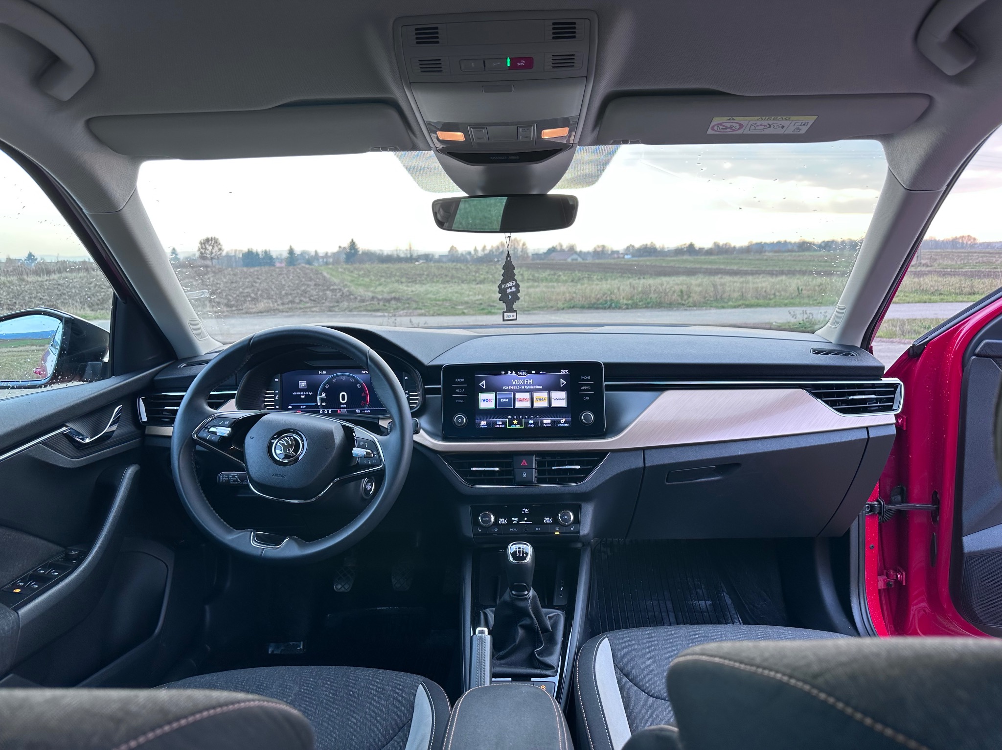 Wnętrze samochodu Skoda z widokiem na deskę rozdzielczą, kierownicę i przednią szybę z zawieszką zapachową. Na zewnątrz widoczny krajobraz polny.
