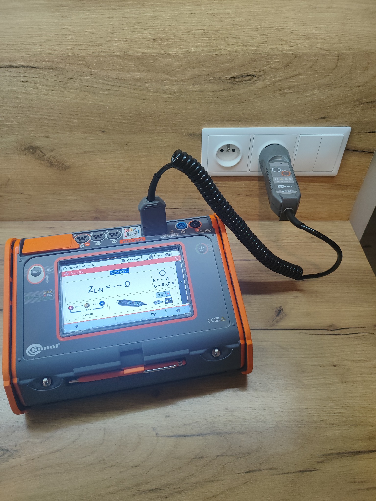 Miernik elektryczny Sonel MPI-540 podłączony do gniazdka, wyświetlający wynik pomiaru impedancji pętli zwarcia. Widoczny przewód pomiarowy i wtyczka adaptera.