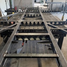 TS WELD TOMASZ SMYTRY - Stalowa konstrukcja ramy spawana na stole spawalniczym z otworami, widoczne ślady spawania i narzędzia w tle warsztatu.