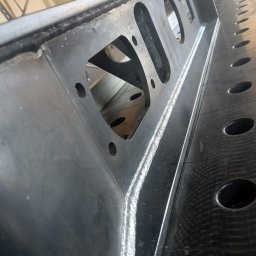 TS WELD TOMASZ SMYTRY - Fragment konstrukcji stalowej z widocznymi spawami i otworami, położonej na perforowanym stole spawalniczym.