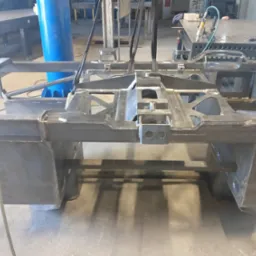 Surowa, spawana konstrukcja stalowa w warsztacie, podwieszona na pasach transportowych, widoczne ślady spawania i obróbki, w tle stoły warsztatowe i narzędzia.