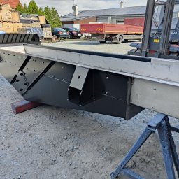 TS WELD TOMASZ SMYTRY - Czarno-srebrna, metalowa konstrukcja skrzyniowa na kozłach warsztatowych, z widocznym w tle wózkiem widłowym i stertą drewnianych palet.