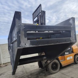 TS WELD TOMASZ SMYTRY - Czarna, surowa konstrukcja stalowa, prawdopodobnie element większej maszyny, podnoszona przez pomarańczowy wózek widłowy na zewnątrz, widoczne ślady spawania i obróbki.