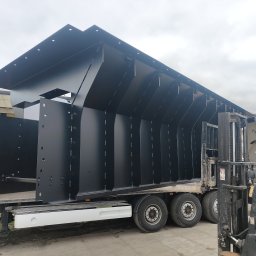 TS WELD TOMASZ SMYTRY - Czarna, stalowa konstrukcja hali, z otworami montażowymi, transportowana na naczepie ciężarówki, widoczny wózek widłowy i operator w czerwonych rękawicach, pochmurne niebo w tle.