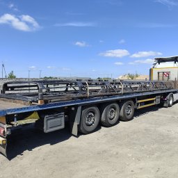TS WELD TOMASZ SMYTRY - Ciężarówka z naczepą przewożąca stalową konstrukcję szkieletową, prawdopodobnie element hali, na tle błękitnego nieba z chmurami i betonowego ogrodzenia.