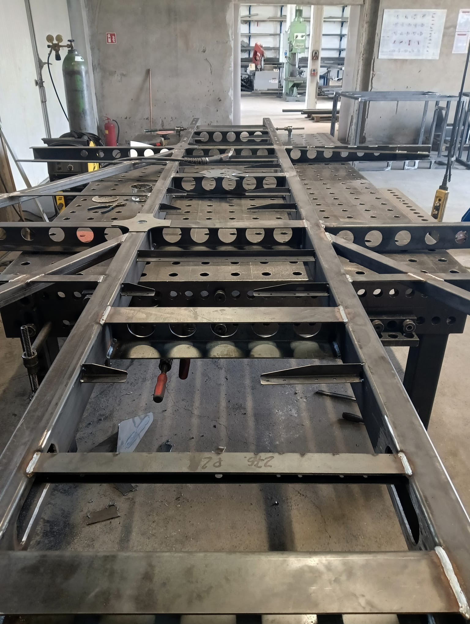 Stalowa konstrukcja ramy spawana na stole spawalniczym z otworami, widoczne ślady spawania i narzędzia w tle warsztatu.