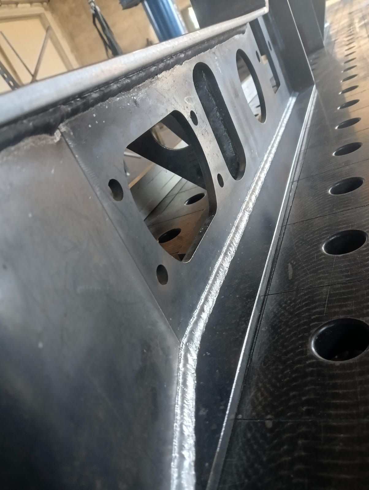 Fragment konstrukcji stalowej z widocznymi spawami i otworami, położonej na perforowanym stole spawalniczym.