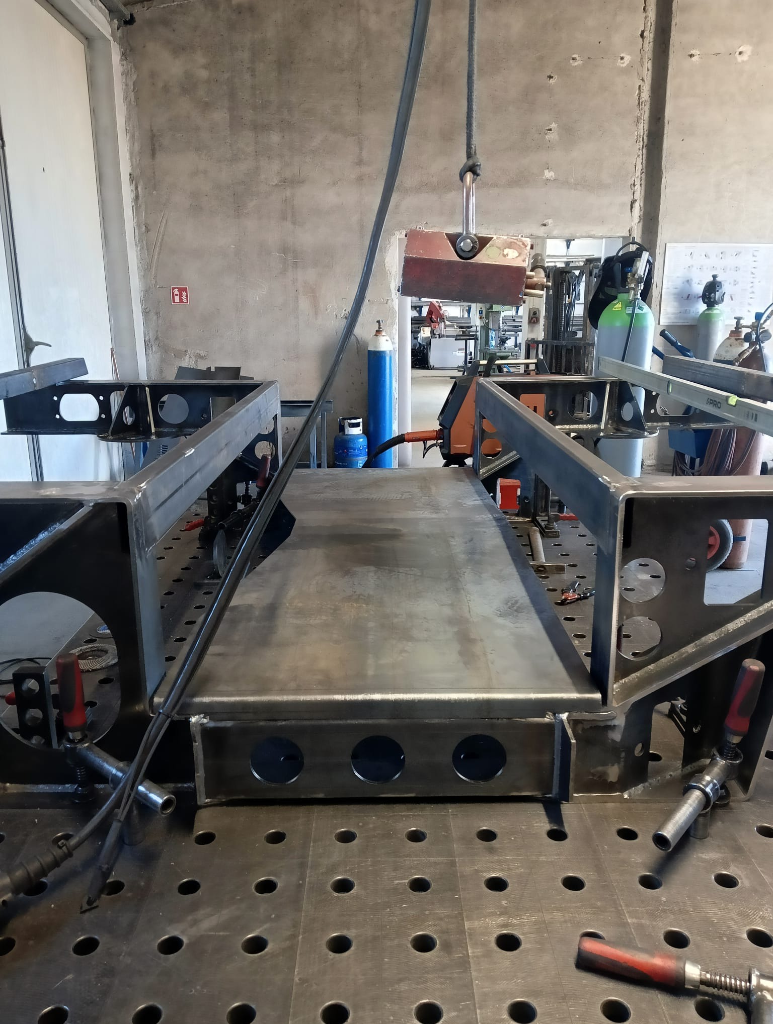 Stalowa konstrukcja spawana na perforowanym stole spawalniczym, widoczne uchwyty spawalnicze, butle z gazem w tle, suwnica nad konstrukcją.