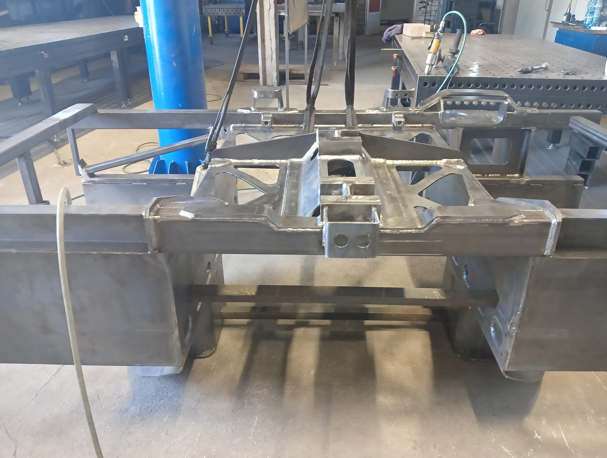 Surowa, spawana konstrukcja stalowa w warsztacie, podwieszona na pasach transportowych, widoczne ślady spawania i obróbki, w tle stoły warsztatowe i narzędzia.