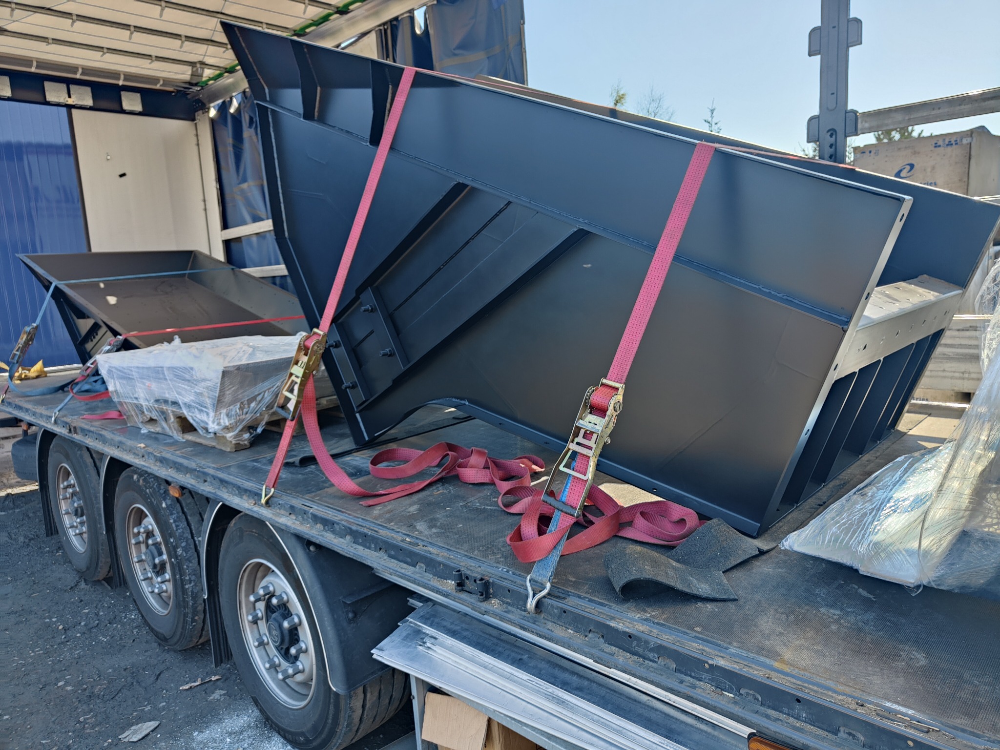 Dwie czarne, metalowe konstrukcje o lejkowatym kształcie, zabezpieczone czerwonymi pasami transportowymi, umieszczone na naczepie ciężarówki podczas transportu.