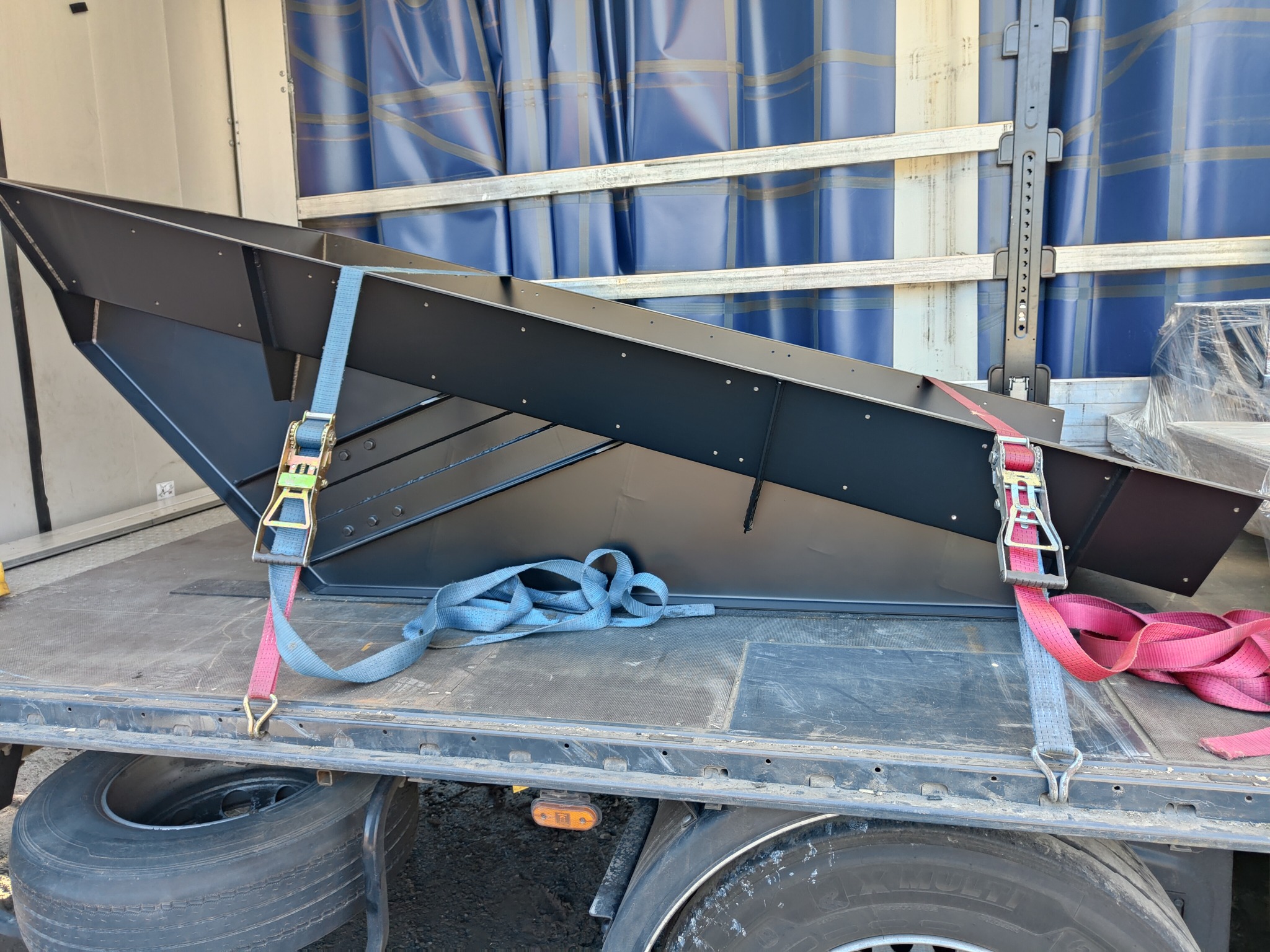 Czarna, metalowa konstrukcja o nietypowym kształcie, zabezpieczona pasami transportowymi na platformie ciężarówki z widocznymi oponami i elementami zawieszenia.