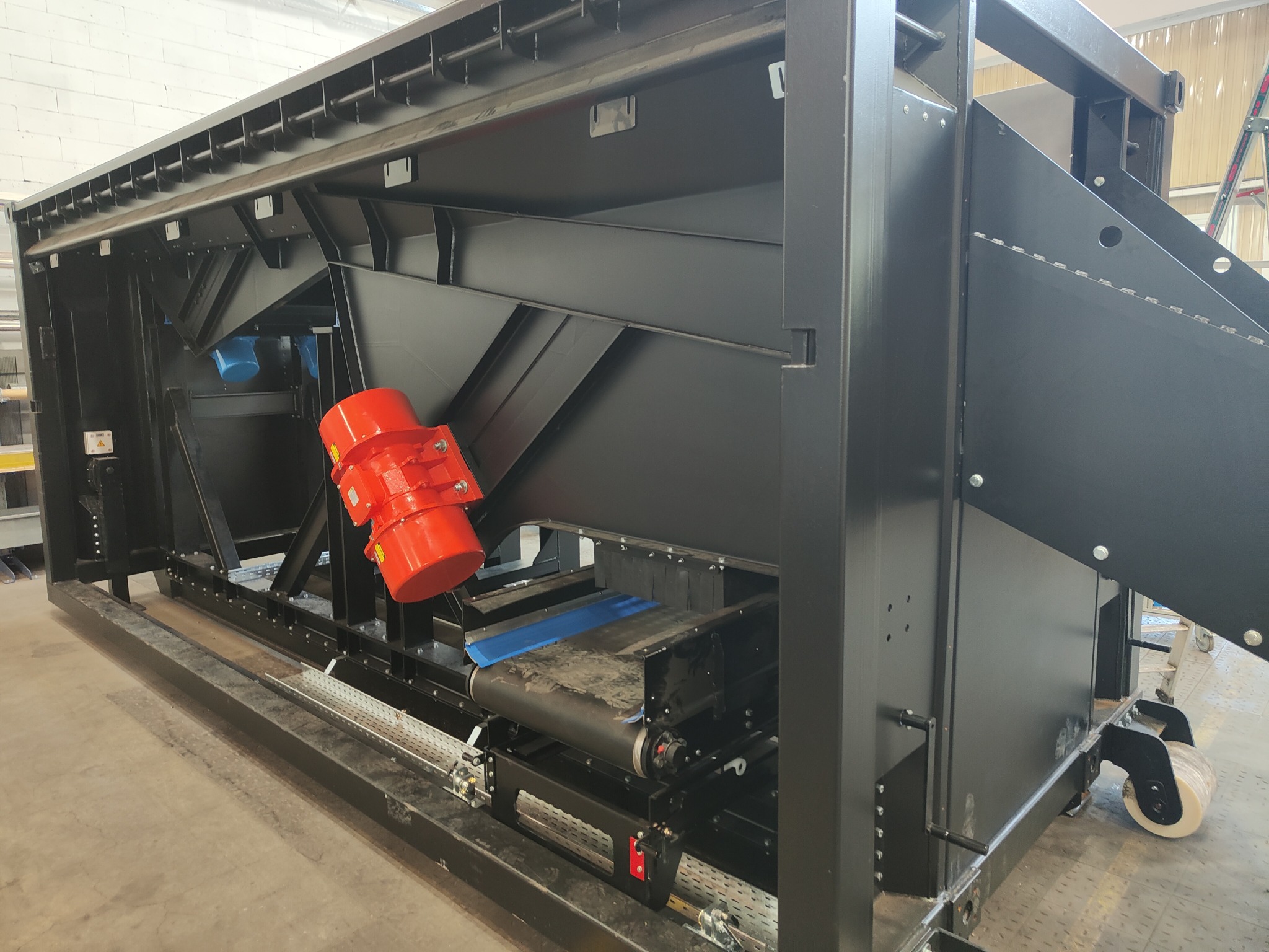 Czarna konstrukcja stalowa z czerwonymi wibratorami i taśmociągiem, widoczne elementy spawane i śruby, całość na kółkach, w tle drabina.