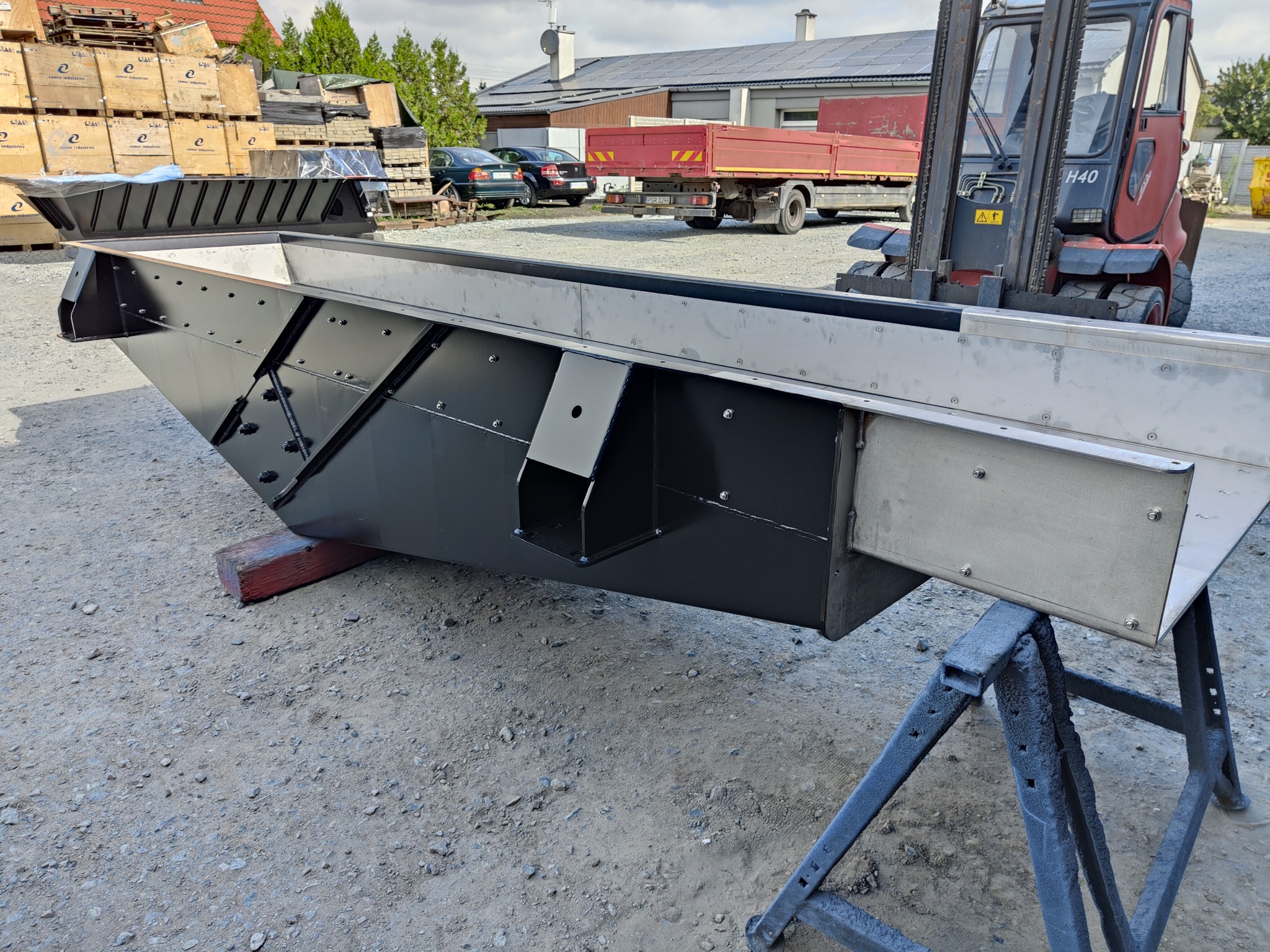 Czarno-srebrna, metalowa konstrukcja skrzyniowa na kozłach warsztatowych, z widocznym w tle wózkiem widłowym i stertą drewnianych palet.