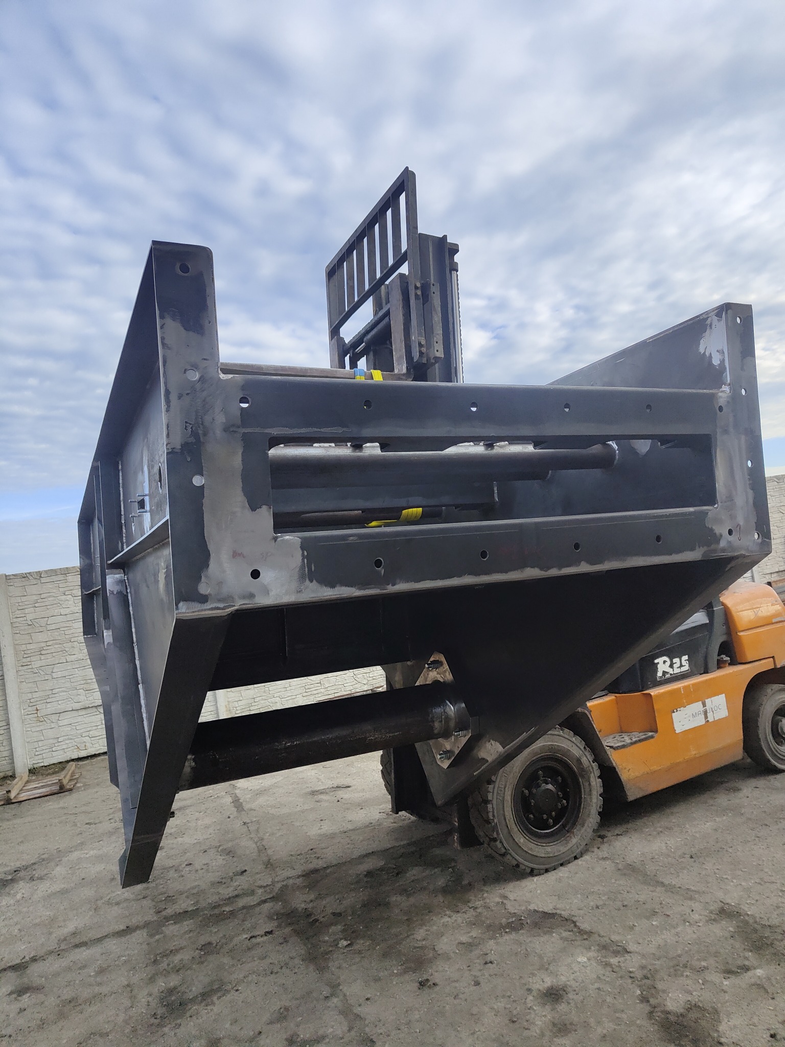 Czarna, surowa konstrukcja stalowa, prawdopodobnie element większej maszyny, podnoszona przez pomarańczowy wózek widłowy na zewnątrz, widoczne ślady spawania i obróbki.