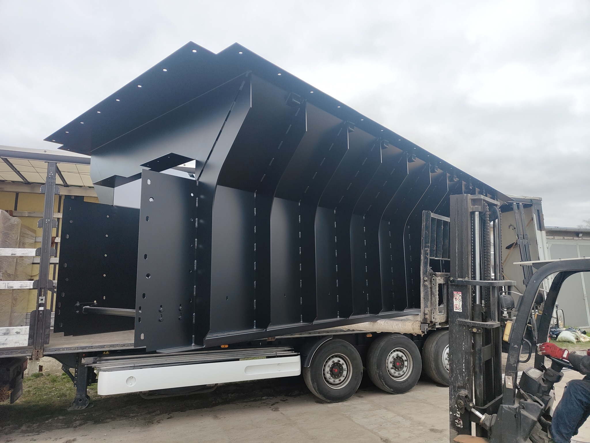 Czarna, stalowa konstrukcja hali, z otworami montażowymi, transportowana na naczepie ciężarówki, widoczny wózek widłowy i operator w czerwonych rękawicach, pochmurne niebo w tle.