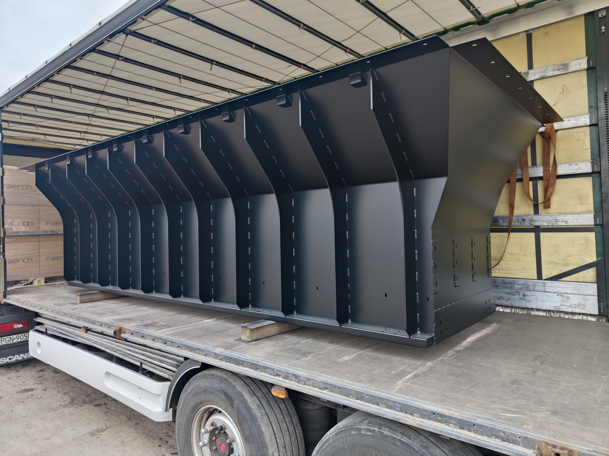 Duża, czarna konstrukcja stalowa z segmentami w kształcie litery V, transportowana na platformie ciężarówki z plandeką.