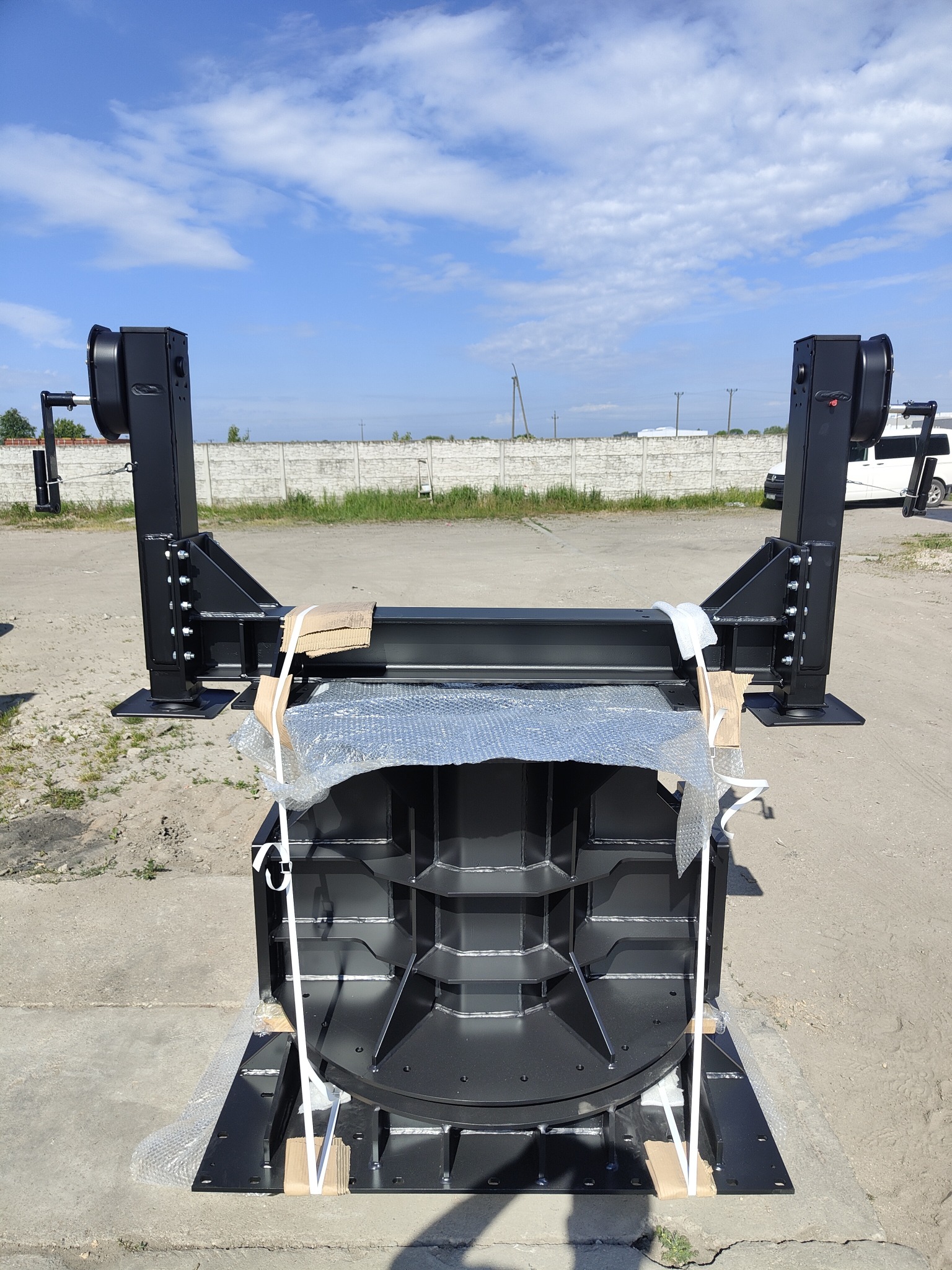 Konstrukcja stalowa w kolorze czarnym, prawdopodobnie element hali, zabezpieczona folią bąbelkową i taśmami, na betonowym podłożu, w tle widoczny mur i błękitne niebo.