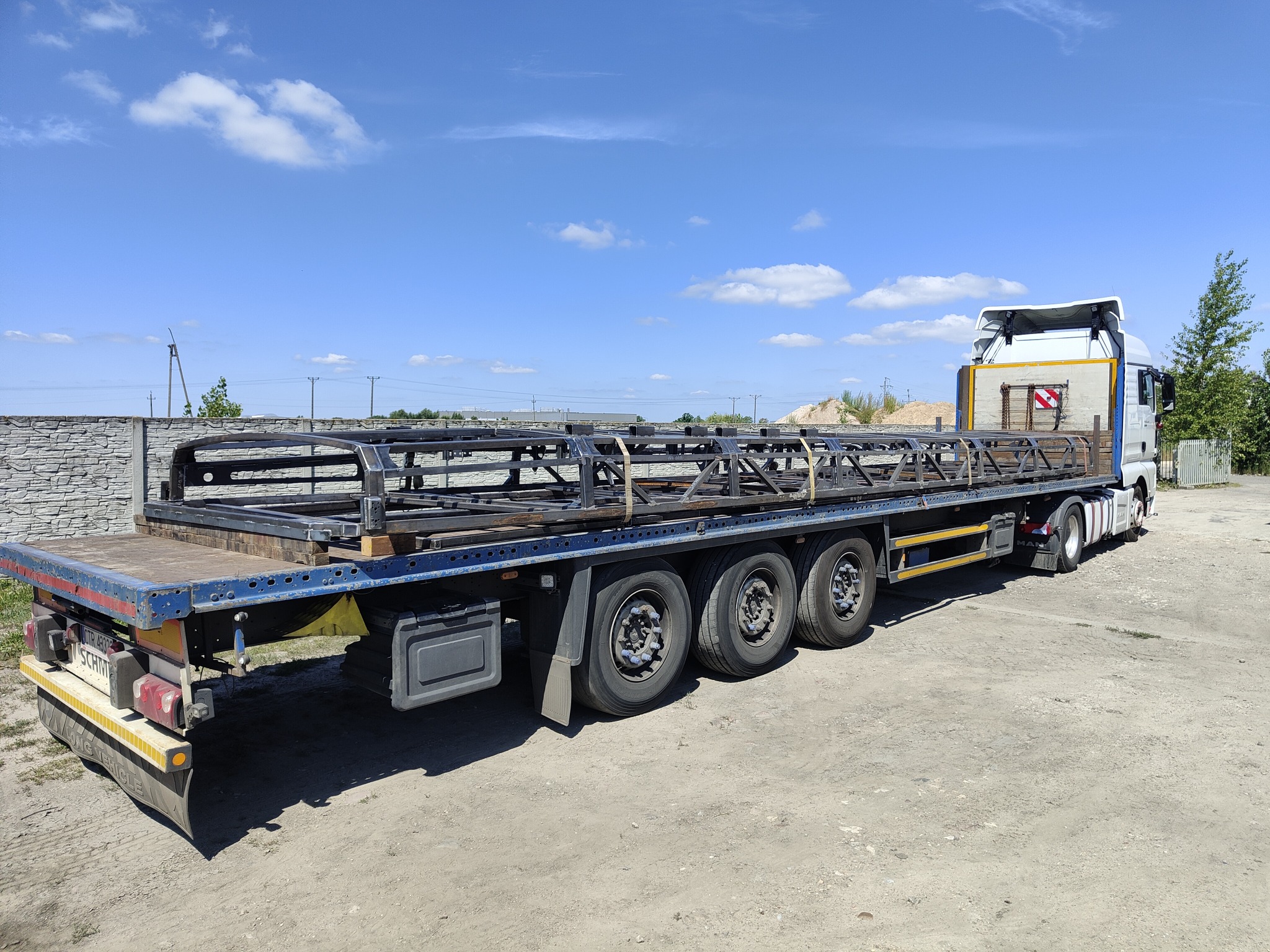 Ciężarówka z naczepą przewożąca stalową konstrukcję szkieletową, prawdopodobnie element hali, na tle błękitnego nieba z chmurami i betonowego ogrodzenia.