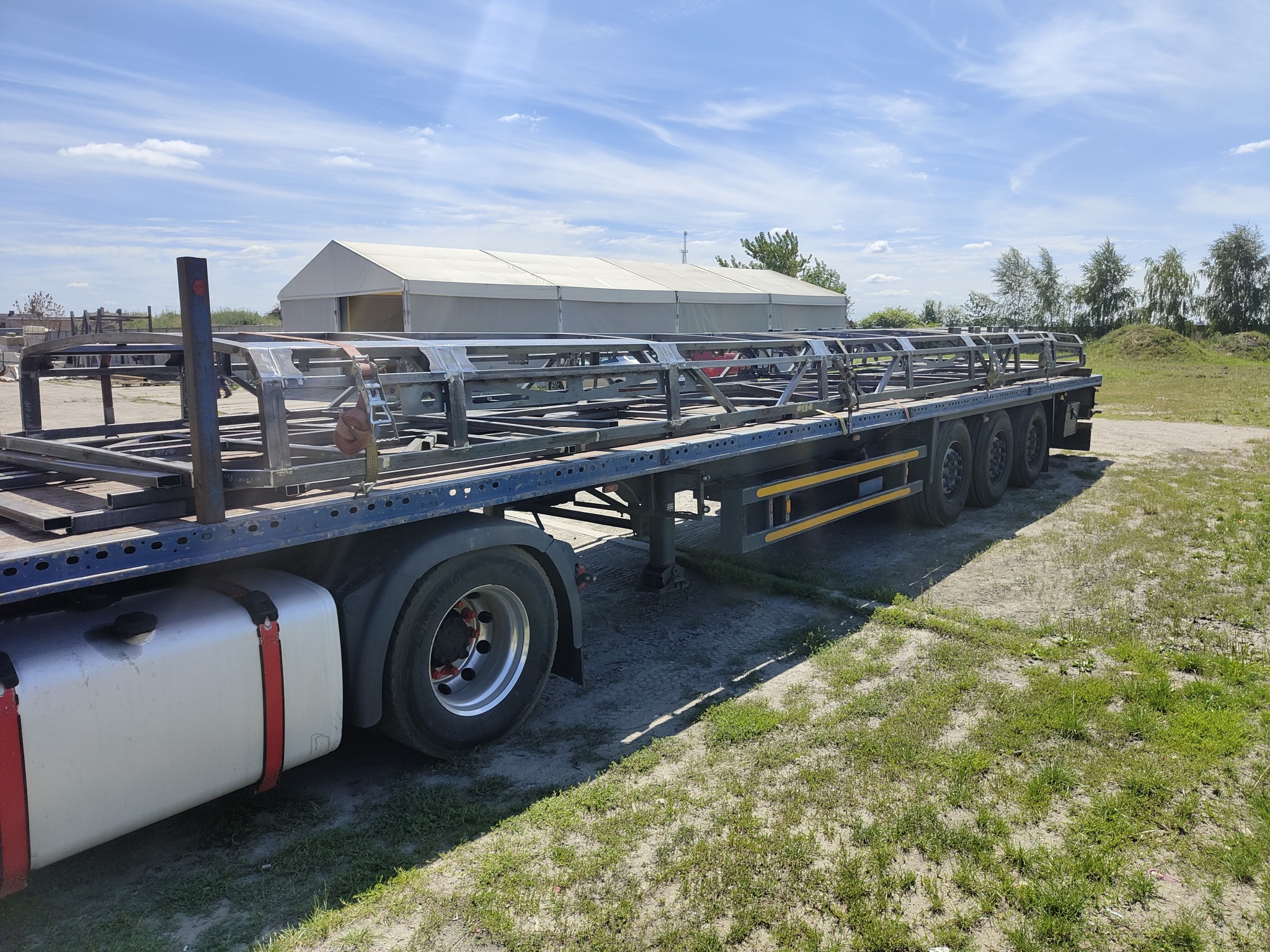 Transport konstrukcji stalowej hali na naczepie ciężarówki w Środzie Wielkopolskiej, widoczna rama i elementy nośne, w tle zarys hali magazynowej.